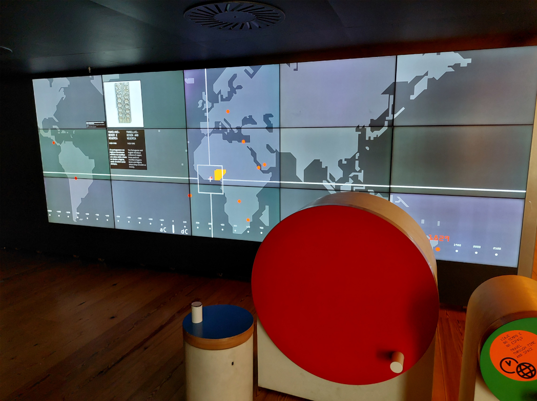 Room containing a big, wide screen, and three colourfol wheels. The screen shows a map of the world, with a cursor on it, pointing at Nigeria.